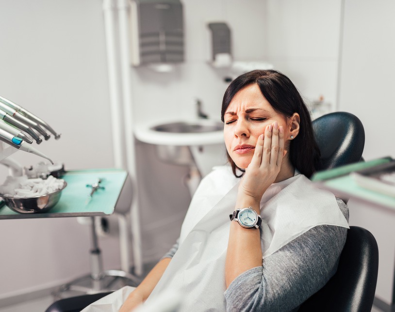 Woman with black hair in dental chair holding hand to cheek in pain