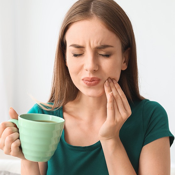 Woman in green t-shirt holding green mug of coffee wincing in pain touching jaw