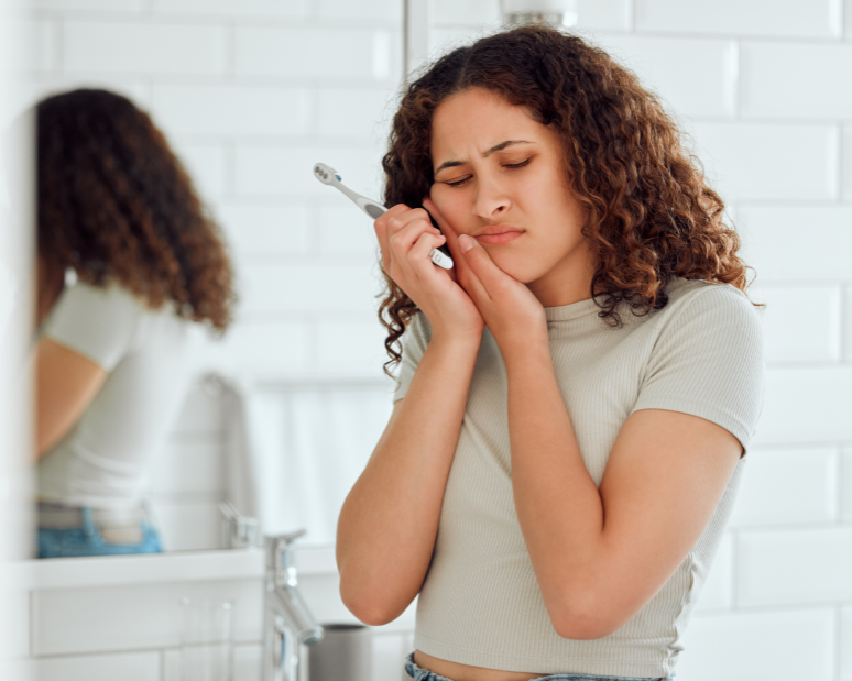 Woman holding jaw with toothbrush in hand, looking uncomfortable